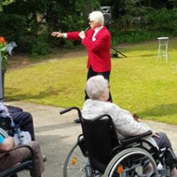 Ein Entertainer im roten Sakko singt im Freien für ein älteres Publikum in Rollstühlen.