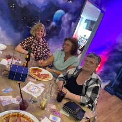 Drei Frauen genießen Pizza an einem Tisch vor einer farbenfrohen Weltraum-Wand, die eine galaktische Atmosphäre schafft.