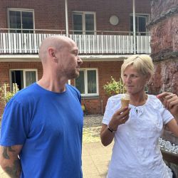 Ein Mann im blauen T-Shirt und eine Frau mit Eis unterhalten sich lebhaft vor einem Backsteingebäude.