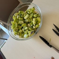 Ein Glasbehälter mit frischem, geschnittenem grünen Salat und Nüssen steht auf einem Tisch neben einer schwarzen Zange.