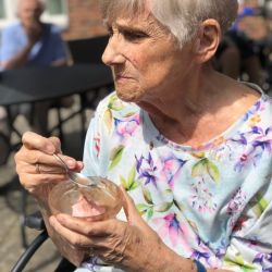 Eine ältere Frau mit blumigem Oberteil genießt draußen im Sonnenschein ein Eis mit einem Löffel in der Hand.