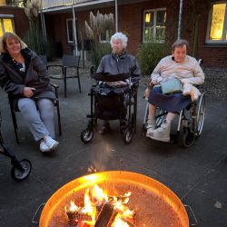 Drei Personen sitzen gemütlich um ein prasselndes Lagerfeuer im Innenhof eines beleuchteten Gebäudes.
