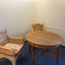 Zwei geflochtene Stühle stehen neben einem runden Holztisch in einem schlichten, gemütlichen Raum.