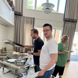Drei Personen bedienen sich an einem Buffet in einem hellen Raum mit hohen Fenstern und eleganten Vorhängen.