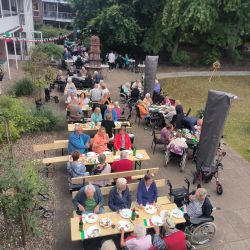 Ältere Menschen sitzen an langen Tischen im Freien und genießen gemeinsam ein geselliges Essen im Grünen.