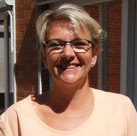 Eine lächelnde Frau mit kurzen blonden Haaren und Brille genießt das sonnige Wetter vor einem Backsteingebäude.