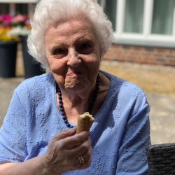 Eine ältere Frau genießt in der Sonne ein Eis, gekleidet in einem blauen Oberteil mit einer Perlenkette.