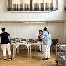 Drei Personen bedienen sich an einem vielfältigen Buffet in einem hellen Raum mit Holzboden und großen Fenstern.