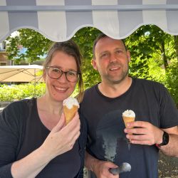 Zwei Personen genießen im Freien unter einem Sonnendach ein Eis und lächeln fröhlich in die Kamera.