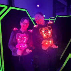Zwei Personen in leuchtenden Lasertag-Westen stehen lächelnd in einer neonbeleuchteten Arena.