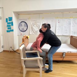 Eine Frau unterstützt eine Person beim Aufstehen von einem Stuhl in einem Schulungsraum mit zahlreichen Plakaten an der Wand.