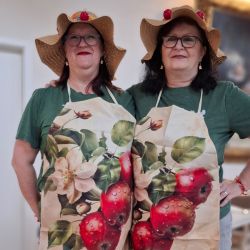 Zwei Frauen tragen Strohhüte und Apfelschürzen, während sie lächelnd nebeneinander auf einem Holzboden stehen.