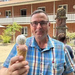 Ein Mann im karierten Hemd genießt ein Eis vor einem Backsteingebäude an einem sonnigen Tag.