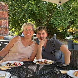 Zwei Personen sitzen entspannt an einem Tisch im Freien und genießen im Sonnenschein ein gemeinsames Essen.