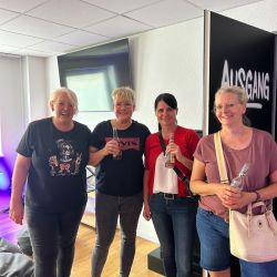 Vier Frauen stehen lächelnd mit Getränken in einem hellen Raum vor einem Schild mit der Aufschrift "Ausgang".