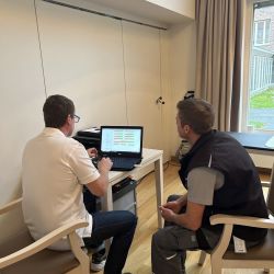 Zwei Männer besprechen am Laptop Daten in einem hellen Raum mit Holzfußboden und großem Fenster.