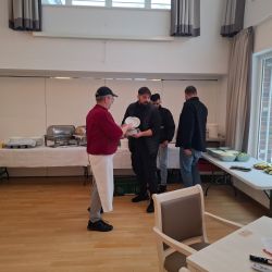 Ein Mann in roter Kochkleidung reicht einem Gast einen Teller, während weitere Gäste am Buffet anstehen.