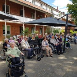 Senioren sitzen entspannt im Freien unter Sonnenschirmen und lauschen einer musikalischen Darbietung in einem Pflegeheim.