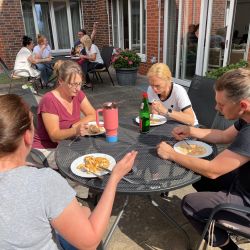 Vier Personen genießen entspannt ein Essen im Freien an einem sonnigen Tag und unterhalten sich angeregt.