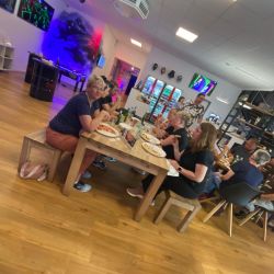 Eine Gruppe von Menschen sitzt in einem modernen Raum und genießt gemeinsam Pizza und Getränke an einem großen Holztisch.