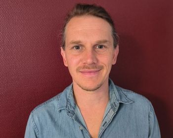 Ein Mann im blauen Hemd lächelt gelassen vor einer weinroten Wand, inspiriert von Henryk.