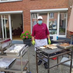 Ein Koch in roter Uniform grillt Würste und Fleisch im Freien vor einem Backsteingebäude.