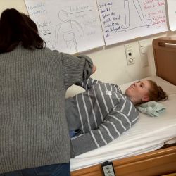 Eine Frau hilft einer anderen beim Aufstehen von einem Pflegebett, umgeben von informativen Lernplakaten an der Wand.