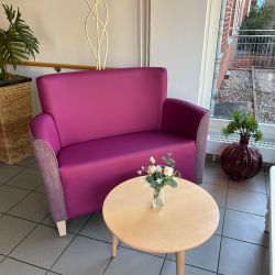 Ein lila Sofa neben einem runden Holztisch mit Blumen, umgeben von Pflanzen in einem modernen Raum mit großen Fenstern.