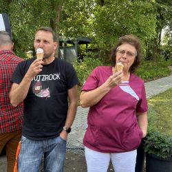 Zwei Personen genießen in entspannter Atmosphäre ein Eis in der Sonne, umgeben von grünen Bäumen und sommerlicher Natur.