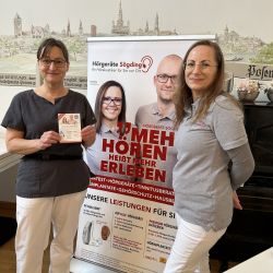 Zwei Frauen stehen lächelnd vor einem Werbebanner für Hörgeräte Sögding in einem Raum mit kunstvoller Wandmalerei.