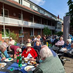 Ältere Menschen genießen ein gemeinsames Mittagessen im Freien vor einem Backsteingebäude an einem sonnigen Tag.