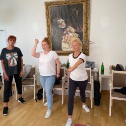 Mehrere Frauen in sportlicher Kleidung machen Dehnübungen in einem hellen Raum vor einem beeindruckenden Gemälde an der Wand.