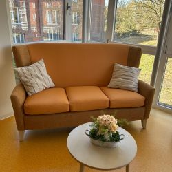 Eine gemütliche, orangefarbene Couch steht vor großen Fenstern, daneben ein runder Tisch mit Blumenarrangement.