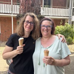 Zwei Frauen stehen lächelnd vor einem Backsteingebäude und genießen an einem sonnigen Tag Eiscreme in Tüten.