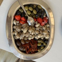 Auf einem Tisch steht eine silberne Platte mit Oliven, gefüllten Paprikas, Pilzen und getrockneten Tomaten bereit zum Servieren.