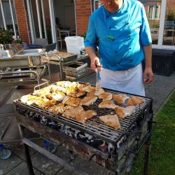 Ein Koch in blauem Hemd grillt mariniertes Fleisch auf einem Holzkohlegrill vor einem Backsteingebäude.