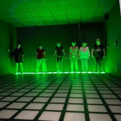 Sechs Personen stehen in einem grün beleuchteten Raum auf kariertem Boden und wirken wie Teil eines futuristischen Szenarios.