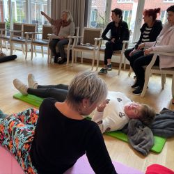 Frauen entspannen auf Yogamatten in einem Raum, während eine Frau vorne Anweisungen gibt.