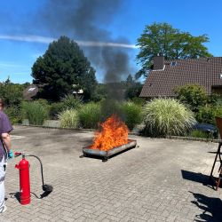 Ein Feuer brennt in einem Metallbehälter auf einem sonnigen Hof, während eine Person mit einem Feuerlöscher bereitsteht.