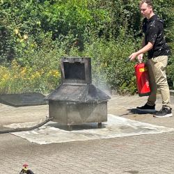 Ein Mann demonstriert im Freien den Einsatz eines Feuerlöschers an einem Metallofen auf einem gepflasterten Platz.
