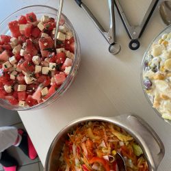 Drei bunte Salate, darunter ein erfrischender Wassermelonen-Feta-Salat, stehen mit Zange und Löffel auf einem Tisch bereit.