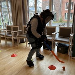 Eine Person in Schutzkleidung spielt mit einem orangenen Hockeyschläger und Ball auf dem Holzboden im Raum.