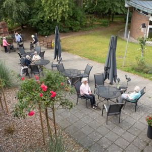 Senioren sitzen entspannt an runden Tischen im Garten eines Heims, umgeben von Blumen und Sonnenschirmen.