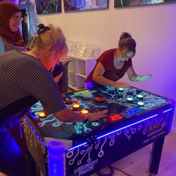 Drei Personen spielen begeistert das interaktive Neon-Lichtspiel "Neuron Race" in einem modernen Spielcenter.