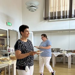 Zwei Personen stehen in einem hellen Raum und genießen Speisen von einem Buffet auf ihren Tellern.