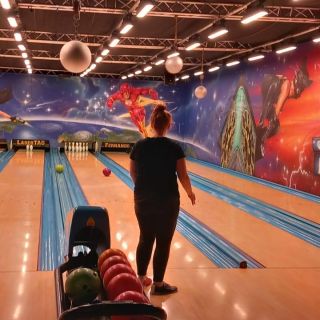 Eine Person wirft eine Bowlingkugel auf der Bahn, während der Spielstand oben auf einem Bildschirm angezeigt wird.