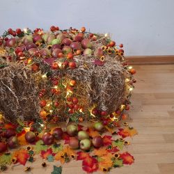 Ein Heuballen, geschmückt mit Lichtern, roten Äpfeln und Herbstblättern, strahlt herbstliche Wärme auf einem Holzboden aus.