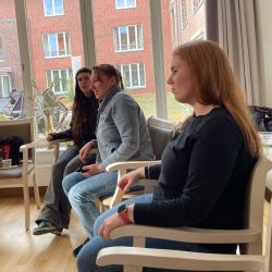 Drei Frauen sitzen in einem hellen Raum und unterhalten sich vor großen Fenstern mit Blick auf ein Backsteingebäude.