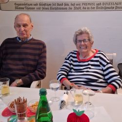 Ein älteres Paar sitzt lächelnd an einem festlich dekorierten Tisch mit Getränken und Snacks in gemütlicher Atmosphäre.