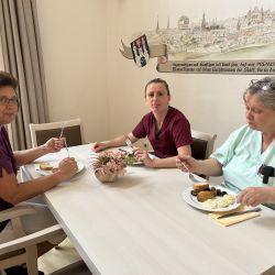 Drei Frauen in Arbeitskleidung genießen ihr Mittagessen an einem hellen Tisch in einem freundlich dekorierten Raum.